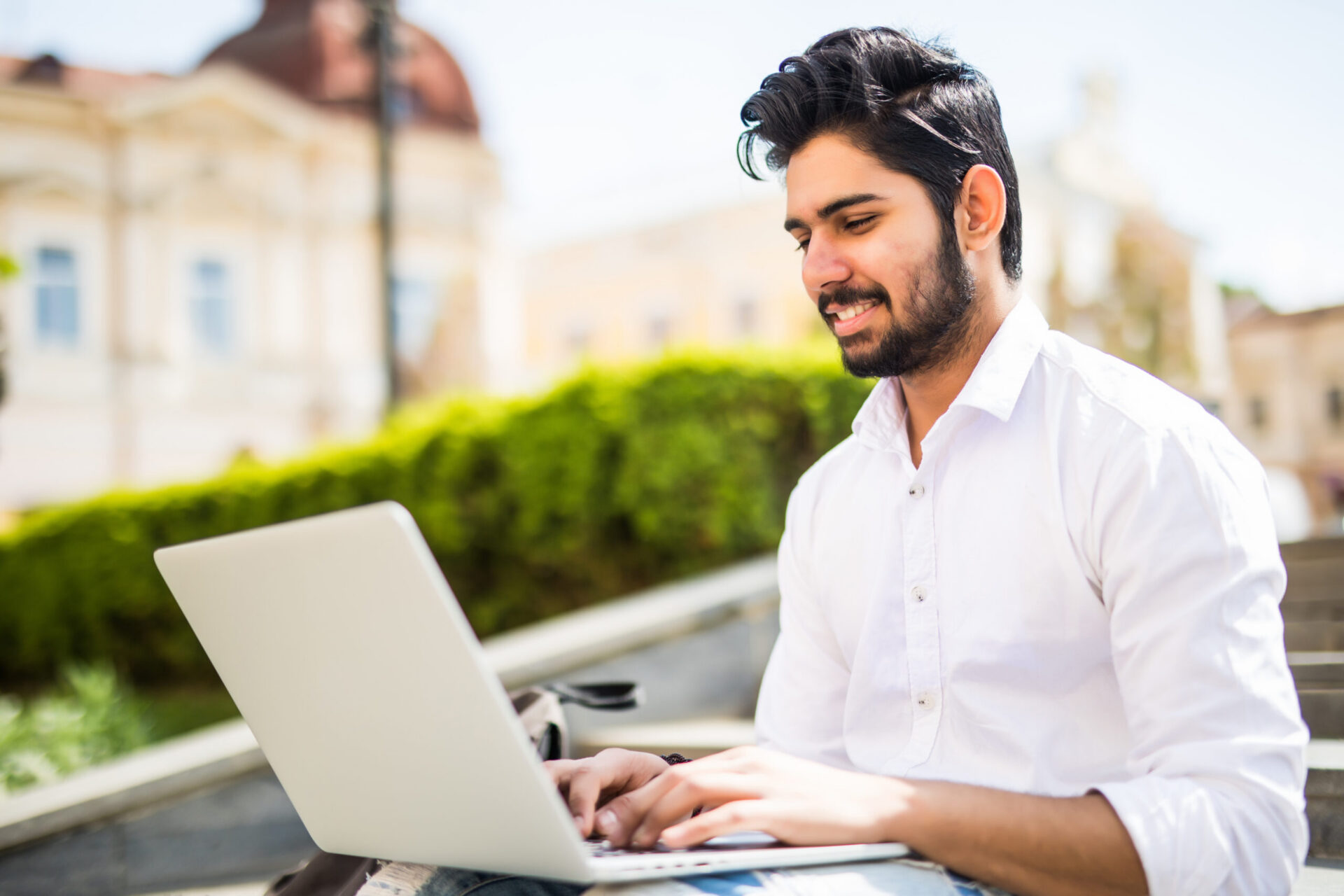 East Indian American College Student sits on stairs on campus, works on laptop computer in the street