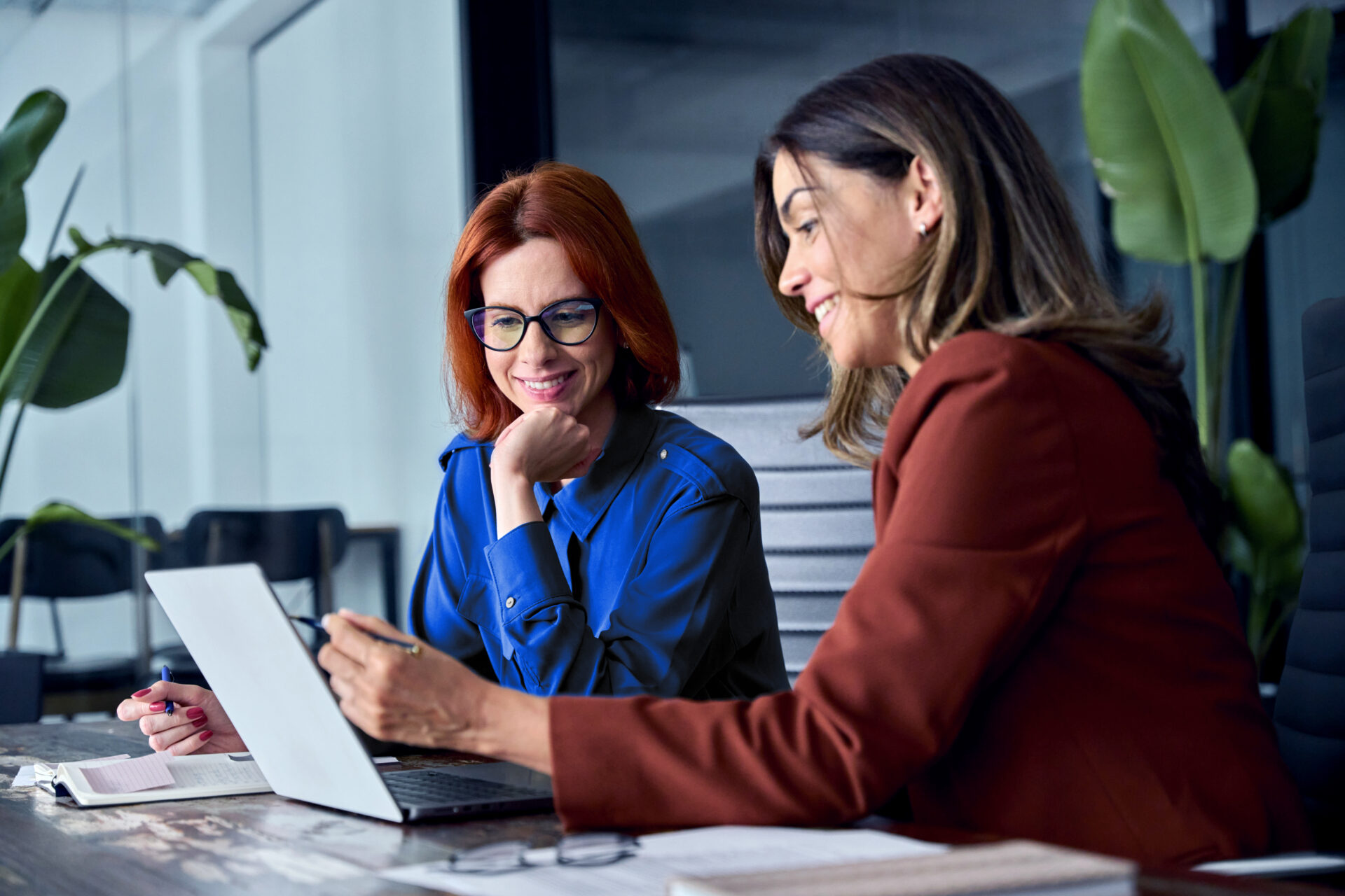 Two professional women review data on a laptop in a modern office, smiling as they collaborate on streamlining financial close processes with AI-powered software. Notebooks and glasses on the table indicate a detailed and analytical discussion, while the setting emphasizes a confident, tech-forward business environment.
